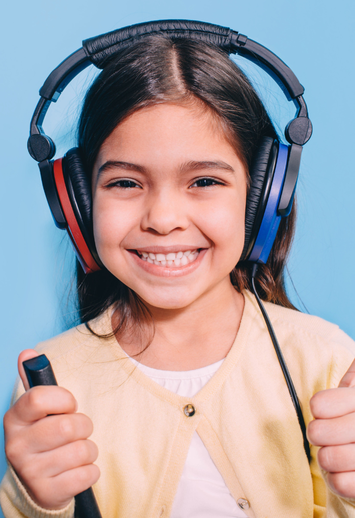 Niña sonriendo con audífonos grandes durante prueba de audición infantil sosteniendo dispositivo de respuesta